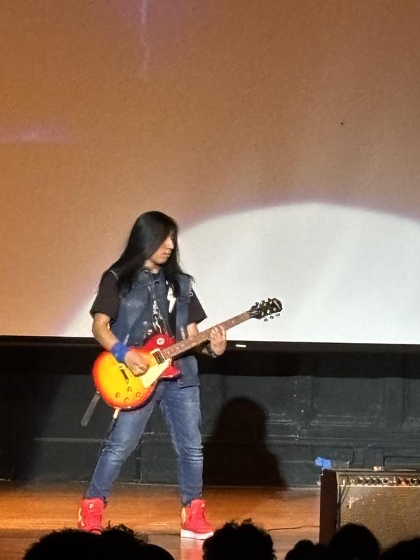A musician with long hair playing an electric guitar on stage, illuminated by a spotlight.