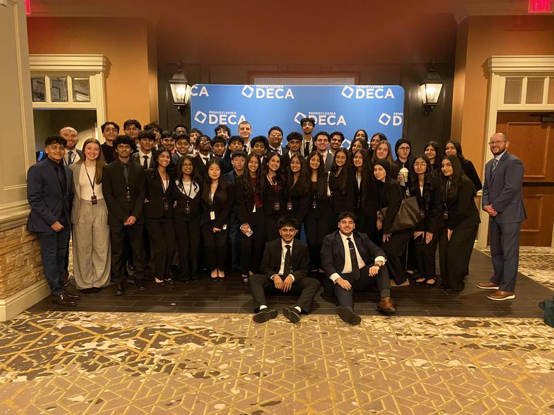 Image of 54 students and their advisor standing in front of a backdrop that says DECA