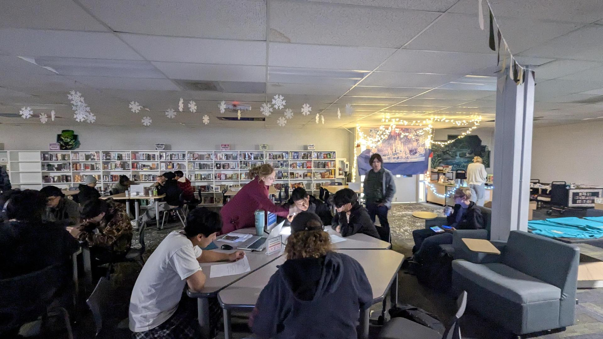 Students working in a library, with bookshelves and holiday decorations visible.