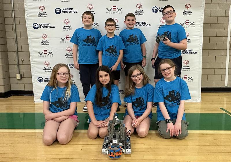 A group of children wearing blue shirts poses with a robot on a gym floor.