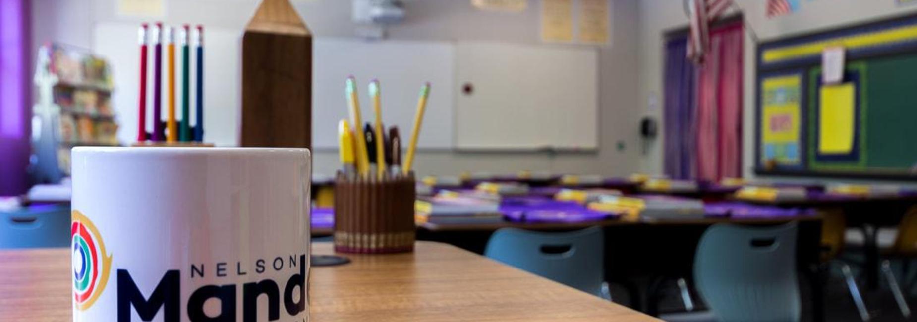 An empty classroom is pictured with a white mug that has the Nelson Mandela Elementary logo on it in the foreground, sitting on a table.