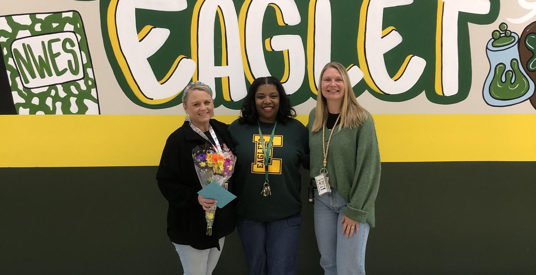 Three women pose in front of a colorful mural with the word 'EAGLET'.