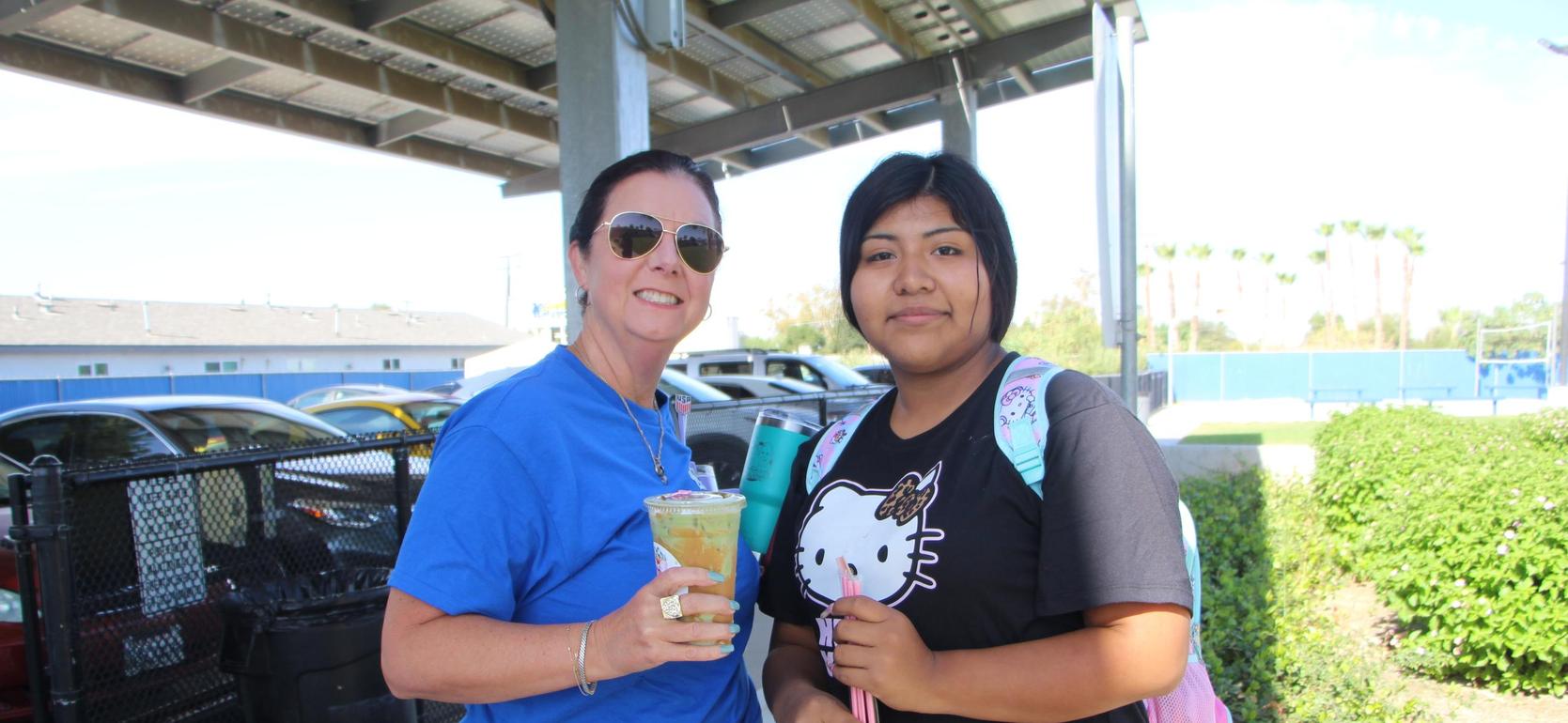 Two women stand outdoors, one holding a colorful drink, with cars and bushes in the background.