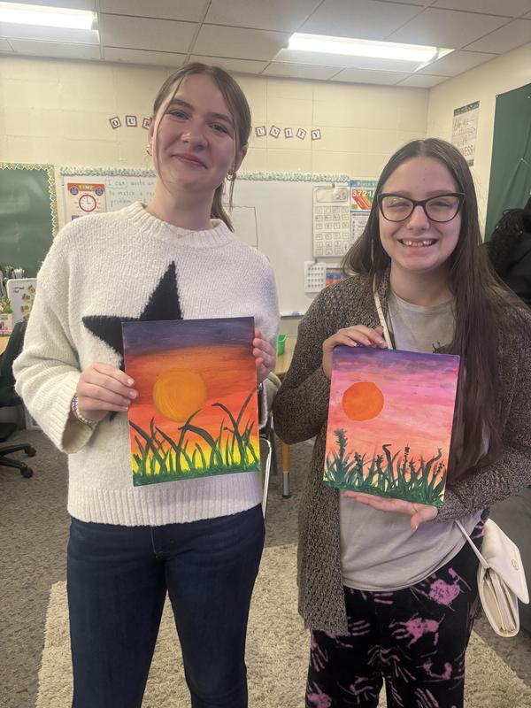 Two girls hold their colorful sunset paintings in a classroom setting.