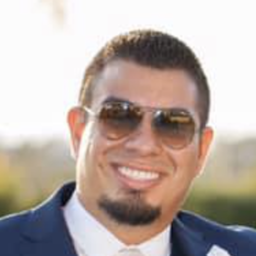 Smiling man in sunglasses and formal attire, outdoors with a blurred background.