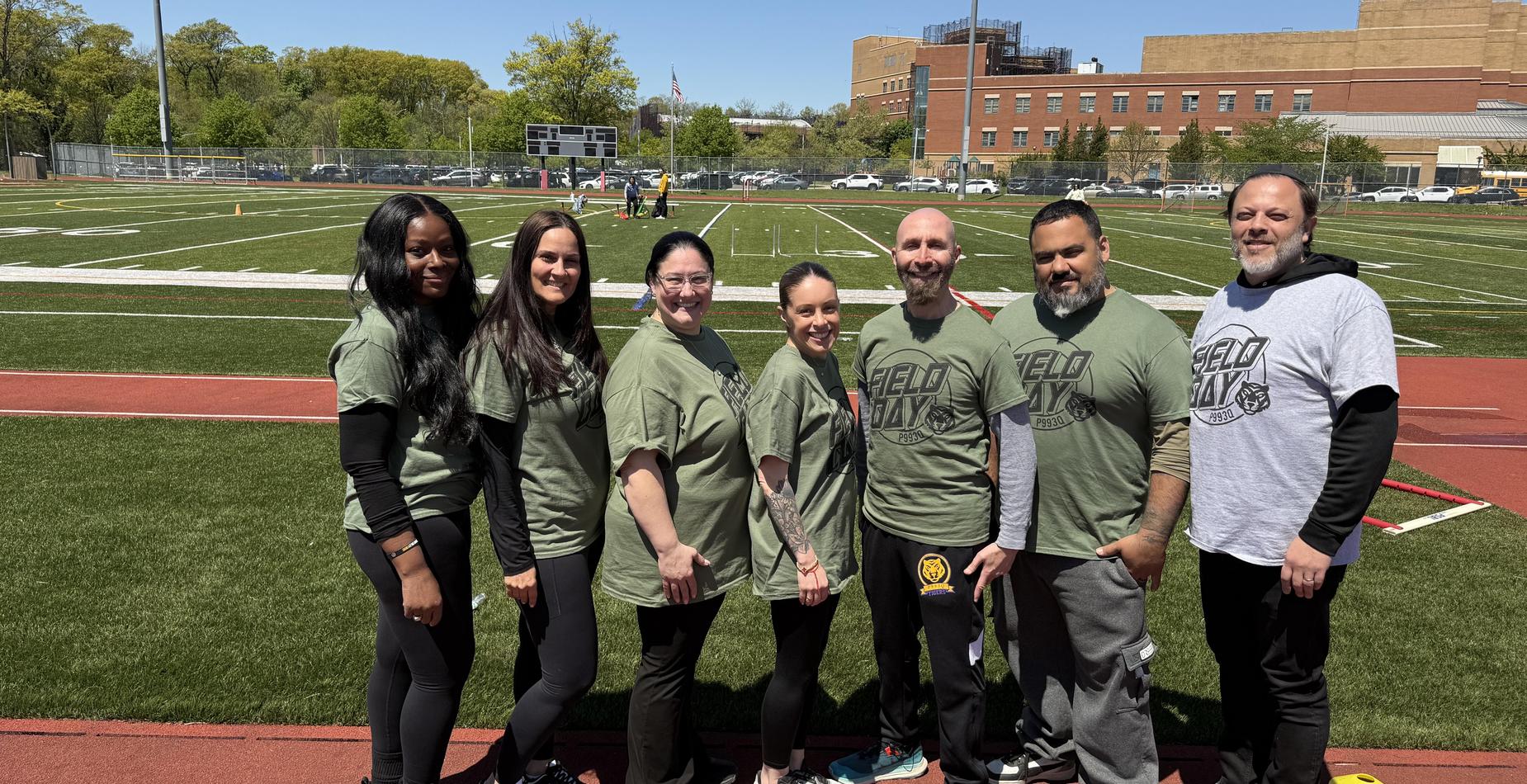 Group of smiling people in green shirts at a sports field.