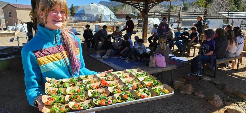 A middle school student at an outdoor learning garden holds a tray full of salad