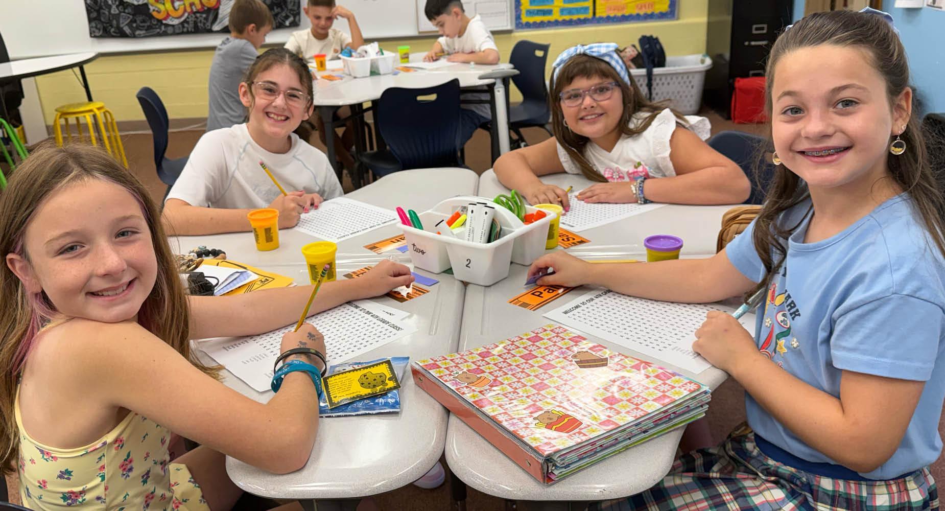 Four children smiling while working at a classroom table with art supplies.