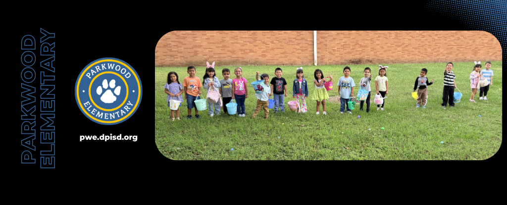 Children standing on grass in front of a brick wall, participating in an event.
