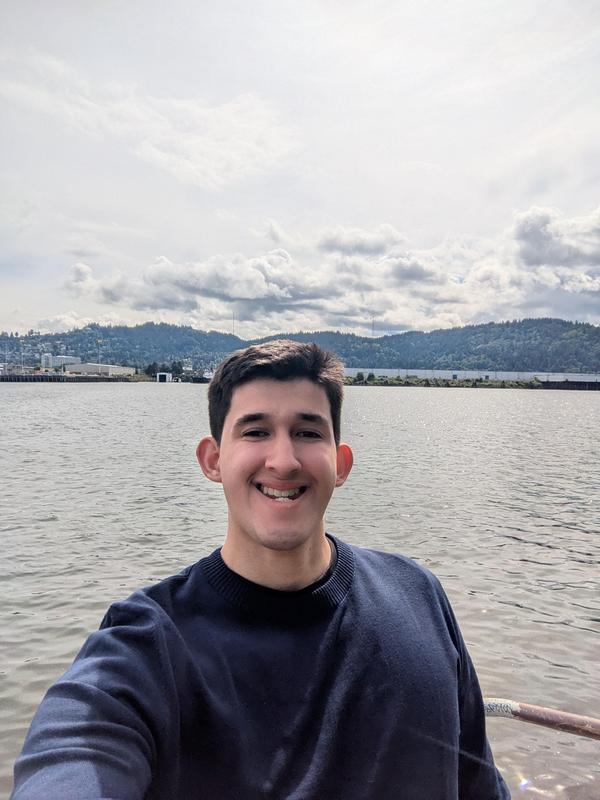 Arco Iris Charter School alum, Zachary Jarvis, stands at a boat railing with water, a town, and hills behind him.