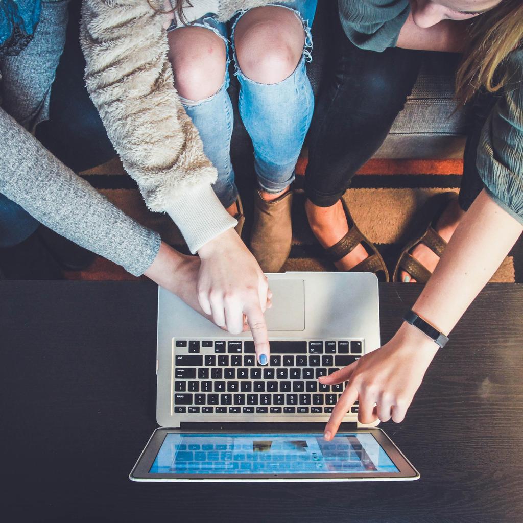 Three people pointing at a laptop from above while seated on a couch.