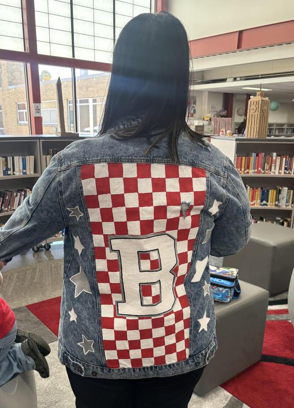 Person wearing a denim jacket with a large red and white checkered B on the back in a library.
