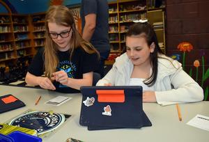 Two girls focus on a game using tablets and pencils in a library.