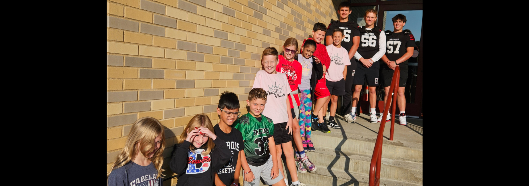 Long line of children in colorful outfits with football players, posing on the stairs outside.