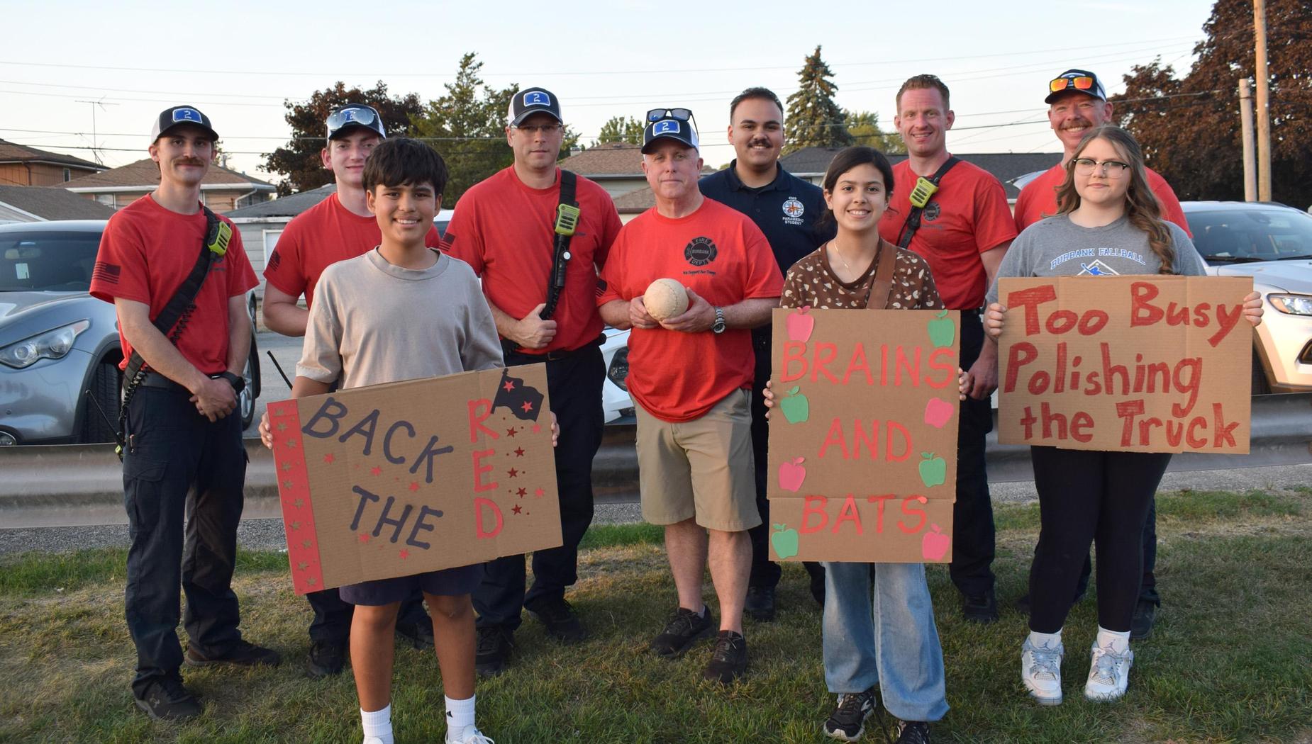 A group of people, including children and adults, pose with signs at a local softball game.