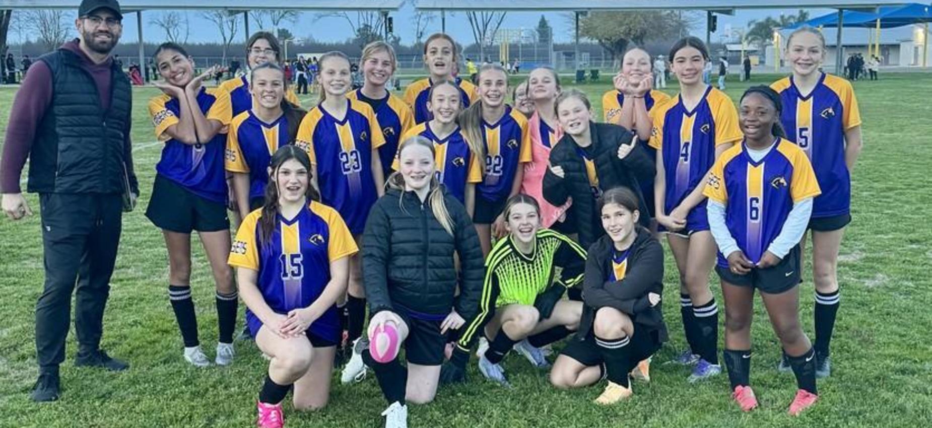 A girls' soccer team poses together on the field, smiling and wearing matching uniforms.
