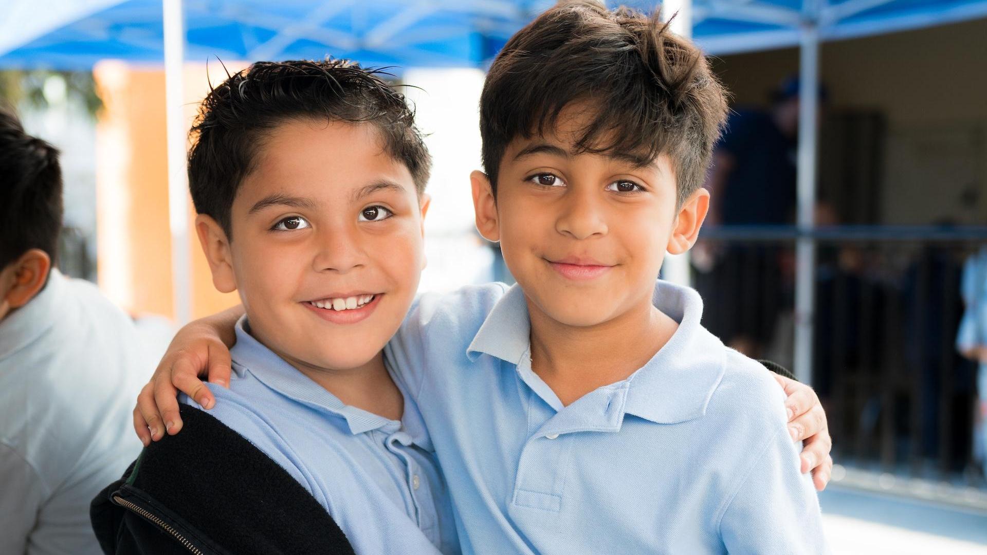 Two boys smiling together, with arms around each other outdoors.