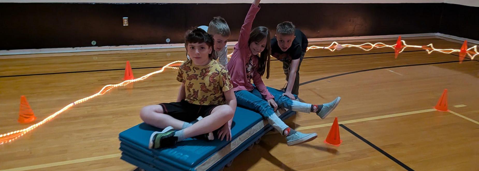 Four children playing on a stack of exercise mats with orange cones and a light trail.
