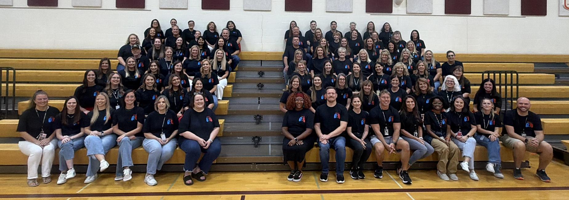 A large group of people in matching black shirts seated on gym bleachers.
