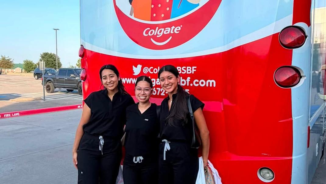 Three women in black uniforms pose in front of a large Colgate-branded bus.