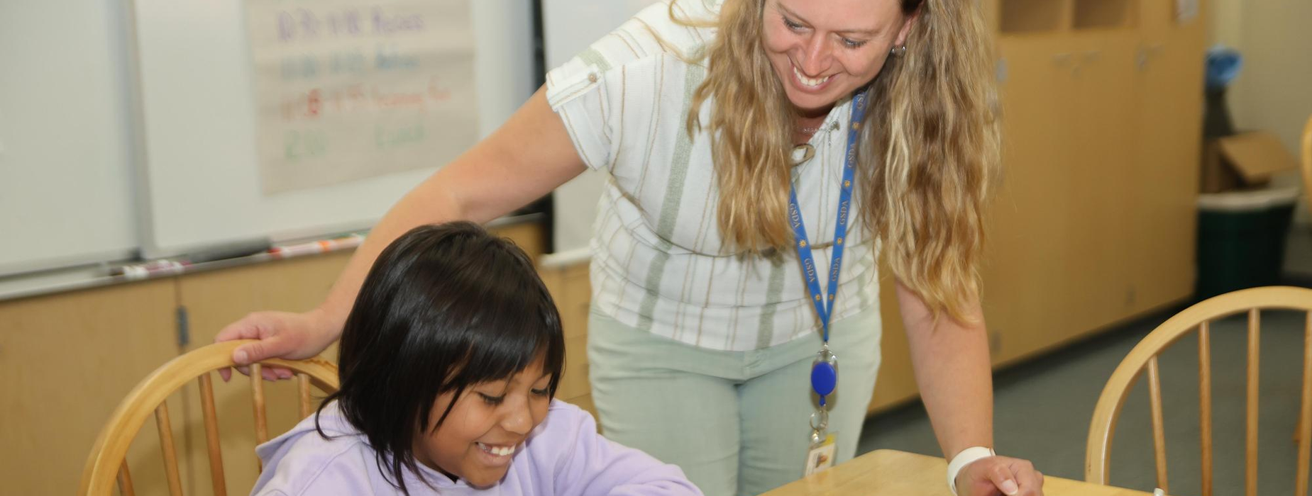 Teacher helping a smiling girl with schoolwork at a table.