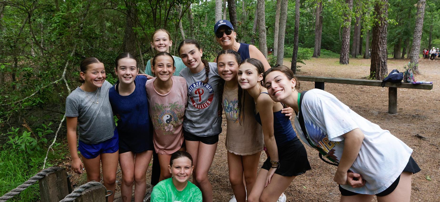 A group of smiling girls and an adult at a wooded outdoor location.