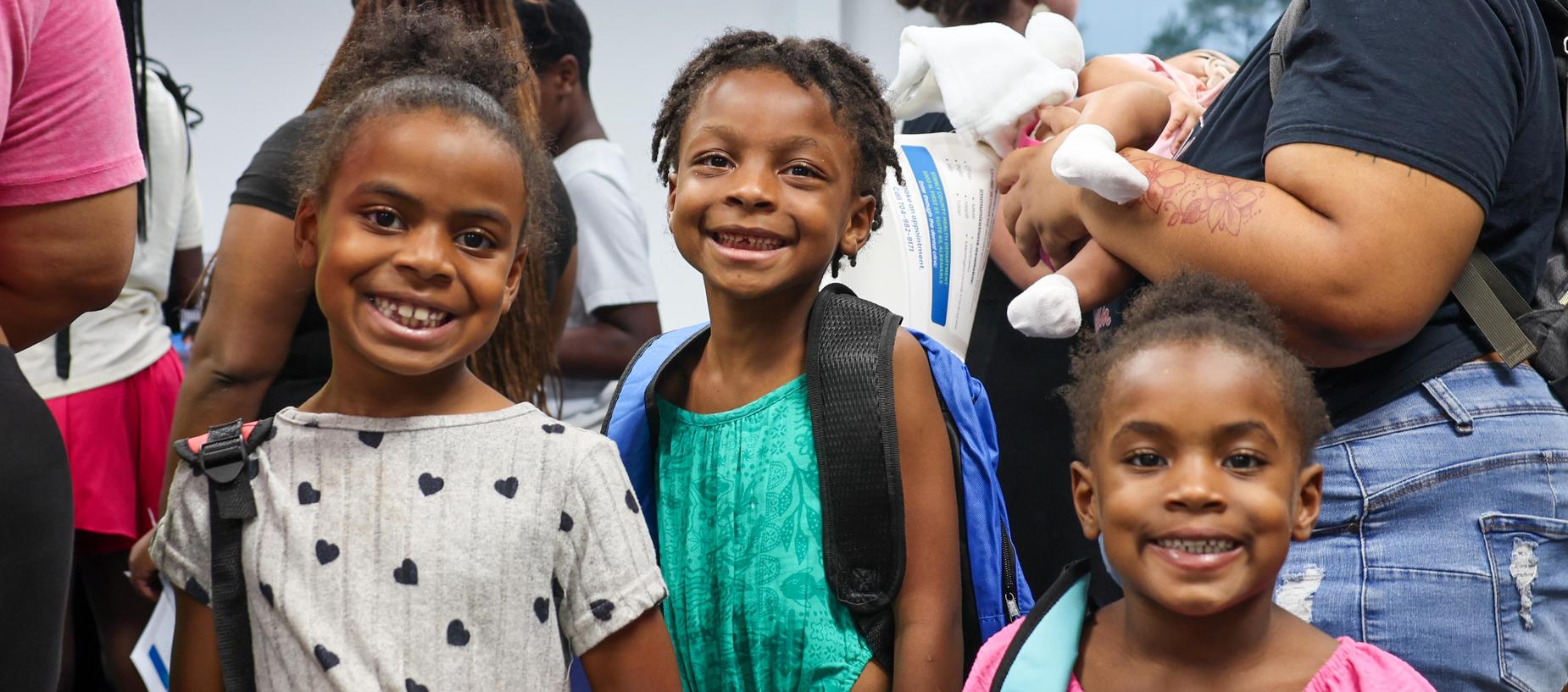 Three smiling children wearing backpacks, standing together in a crowd.