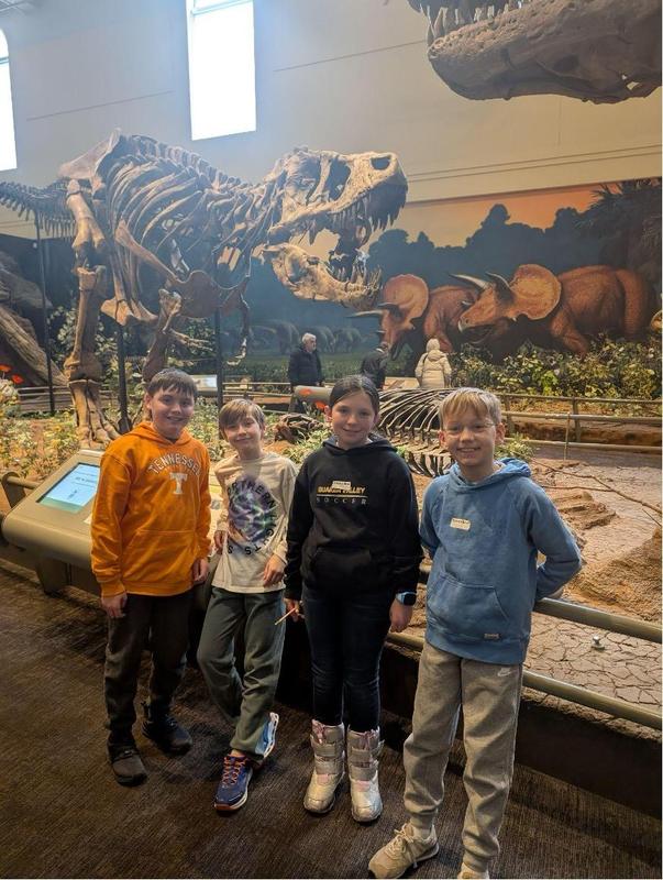 Four smiling students leaning against a dinosaur exhibit at the museum.