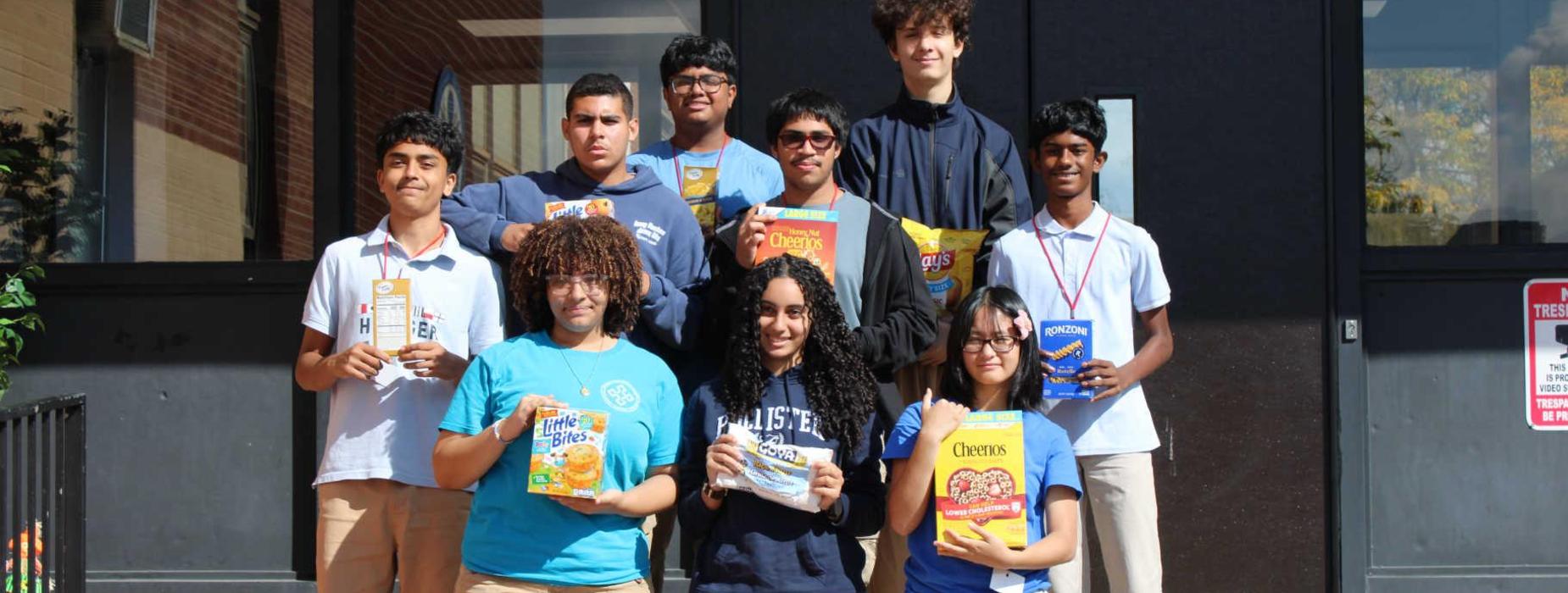 Group of students holding cereal boxes, standing at a school entrance.