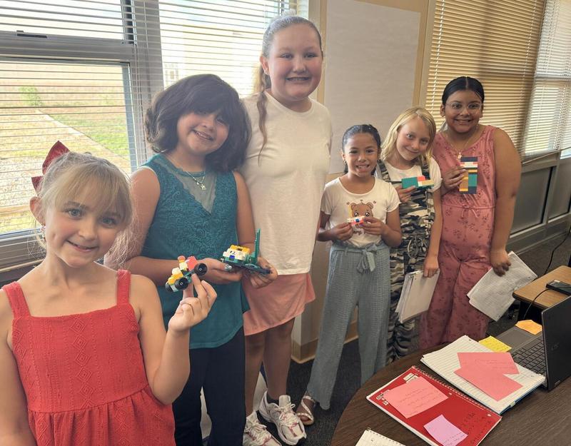 Six smiling students standing in a row show off their LEGO creations inside a school classroom.