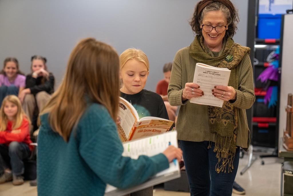 Teacher is reading the script with two other students participating.  There are students in the background watching.
