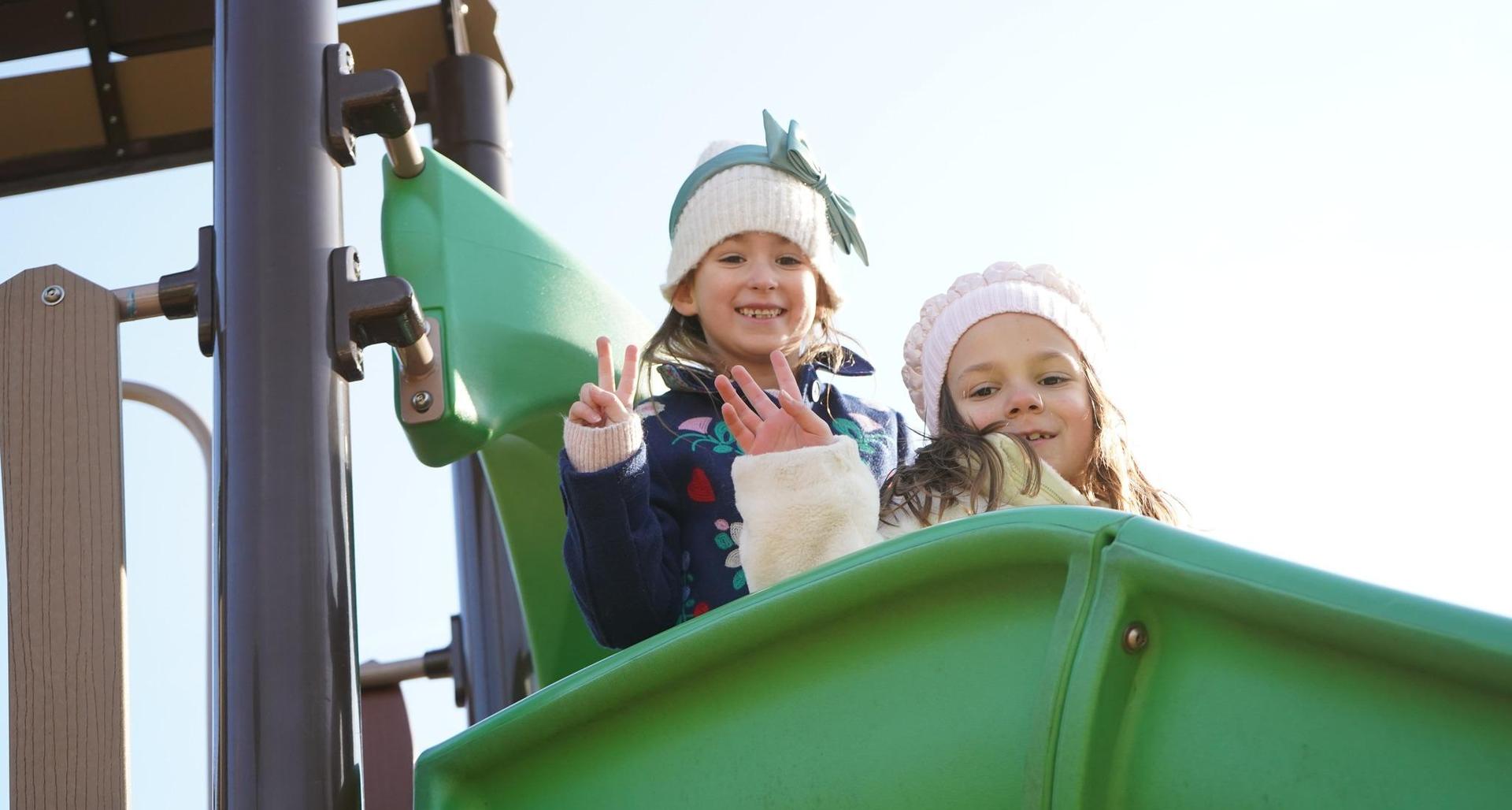 Two girls smile and wave from a green slide at a playground.