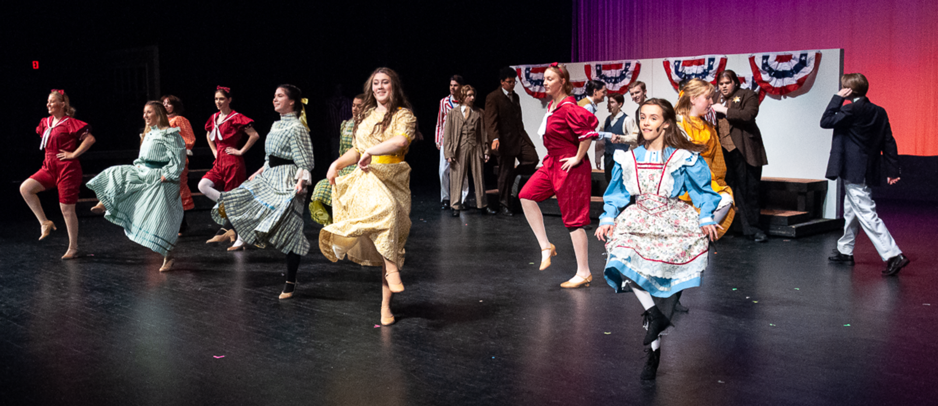 The Music Man production with women dressed in dresses while dancing on the stage with red, white and blue banners and men in suits in the background