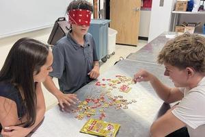 Three students work together around a metal table to assemble a puzzle, with one blindfolded person placing pieces.