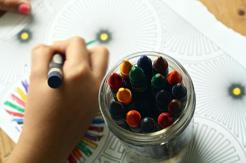 A child's hand holding a crayon, coloring next to a glass jar of crayons.