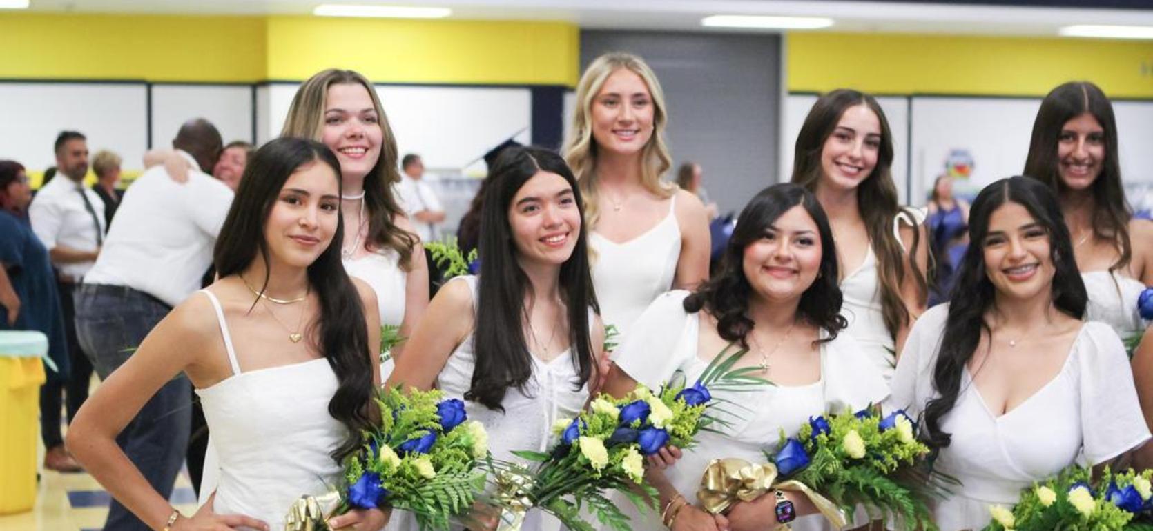 Group of young women in white dresses, smiling with bouquets of flowers in a hall.
