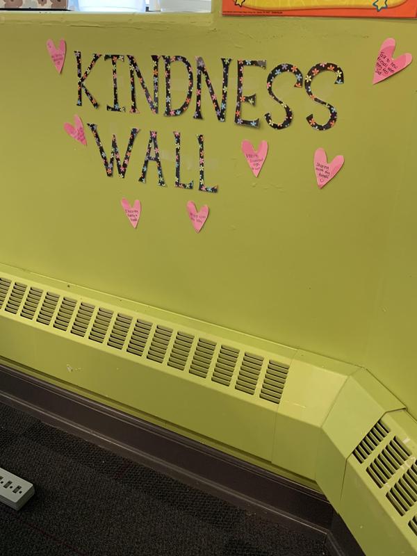 Image shows a green wall with the text "Kindness Wall", surrounded by hearts