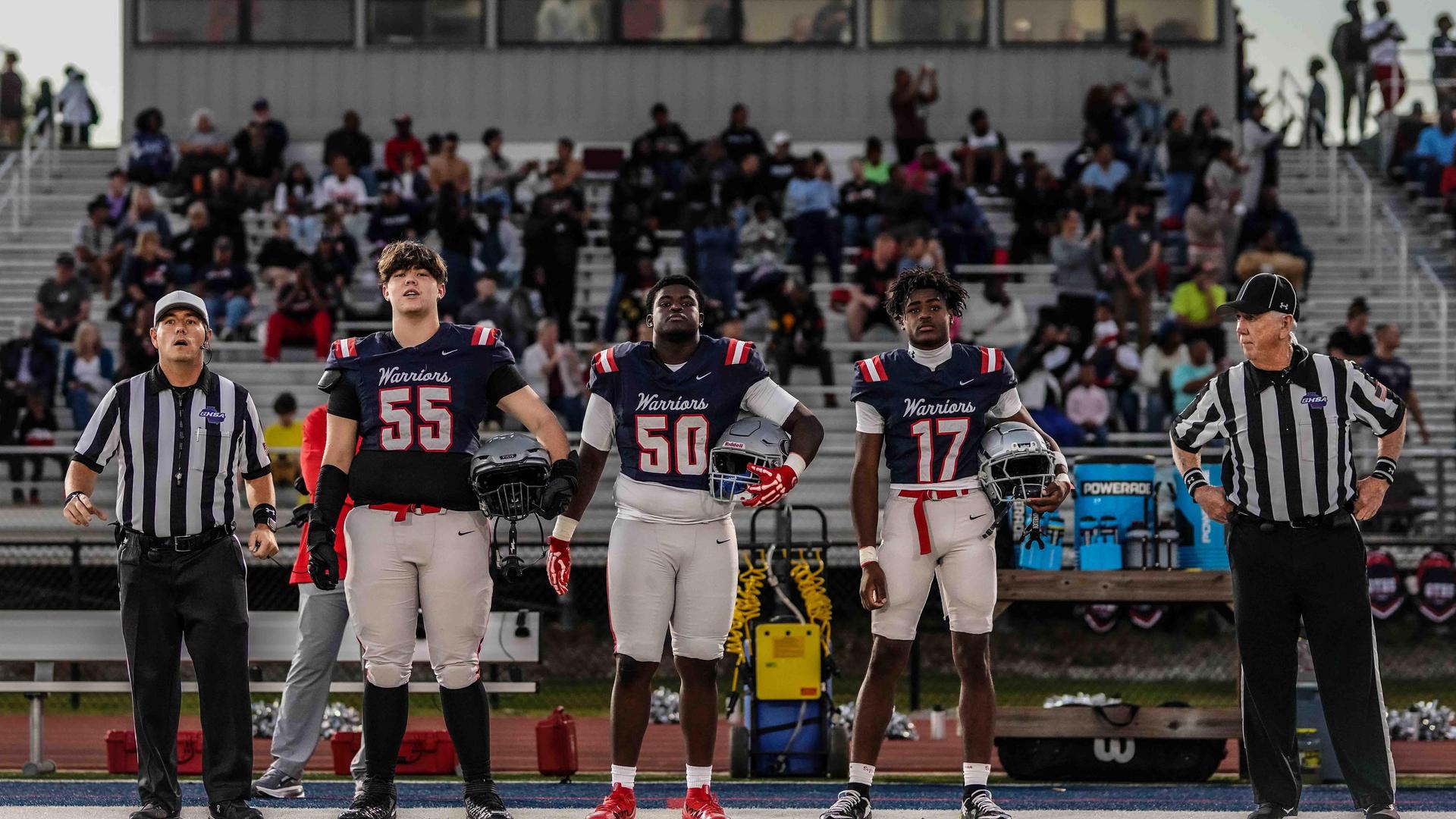 Three football players standing together in uniform, holding helmets, with referees beside them.