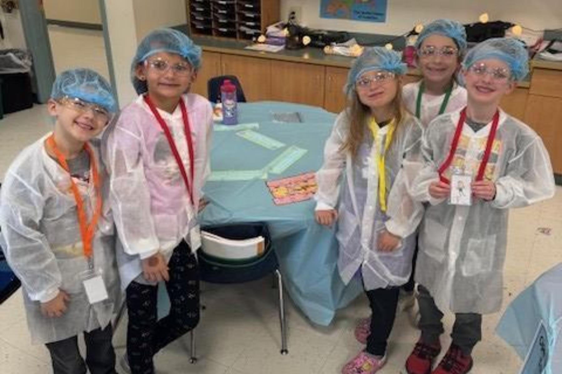 Five children wearing lab coats and hats, smiling together in a classroom setting.