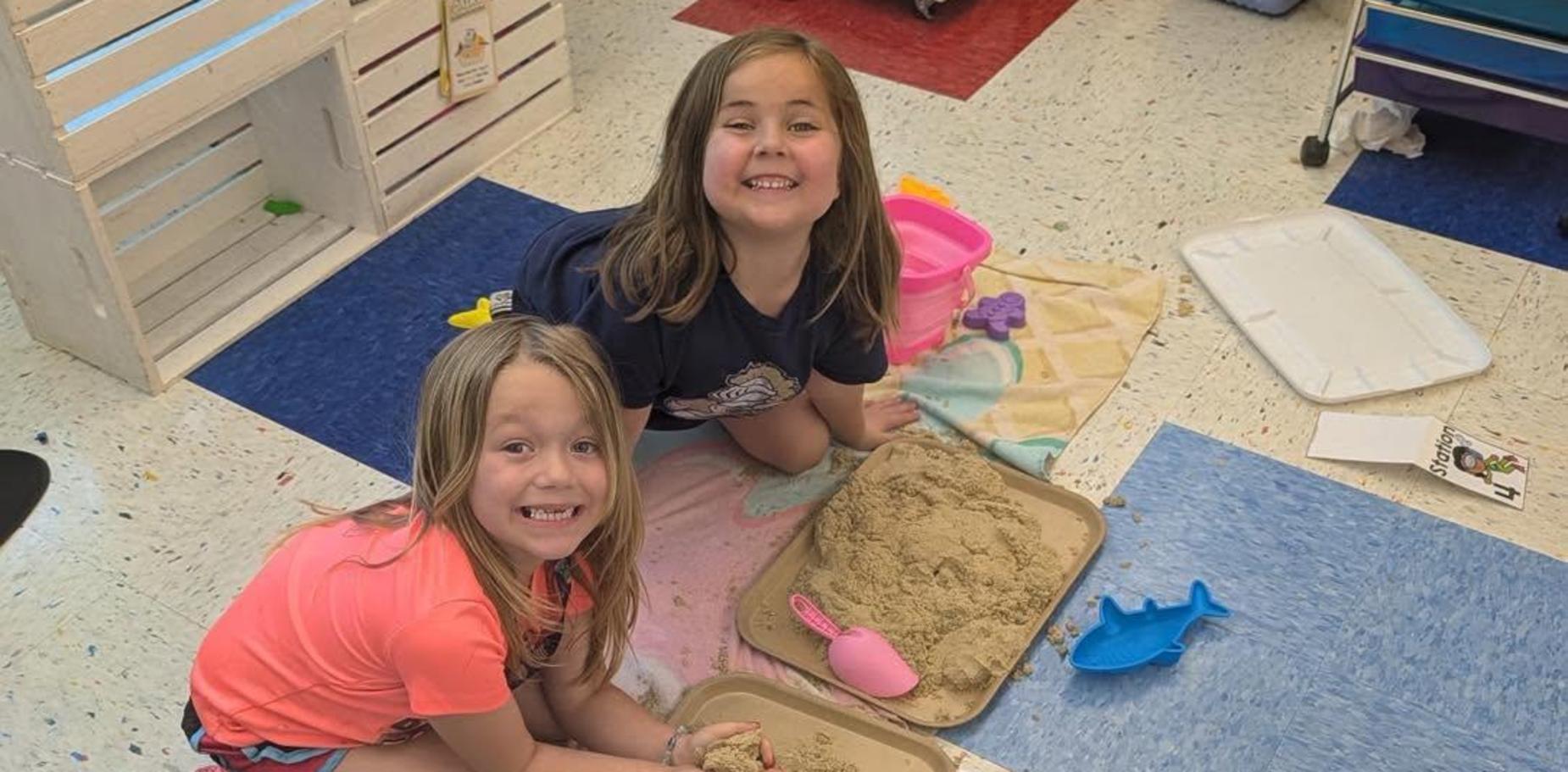 Two children play with sand on trays in a colorful classroom setting.