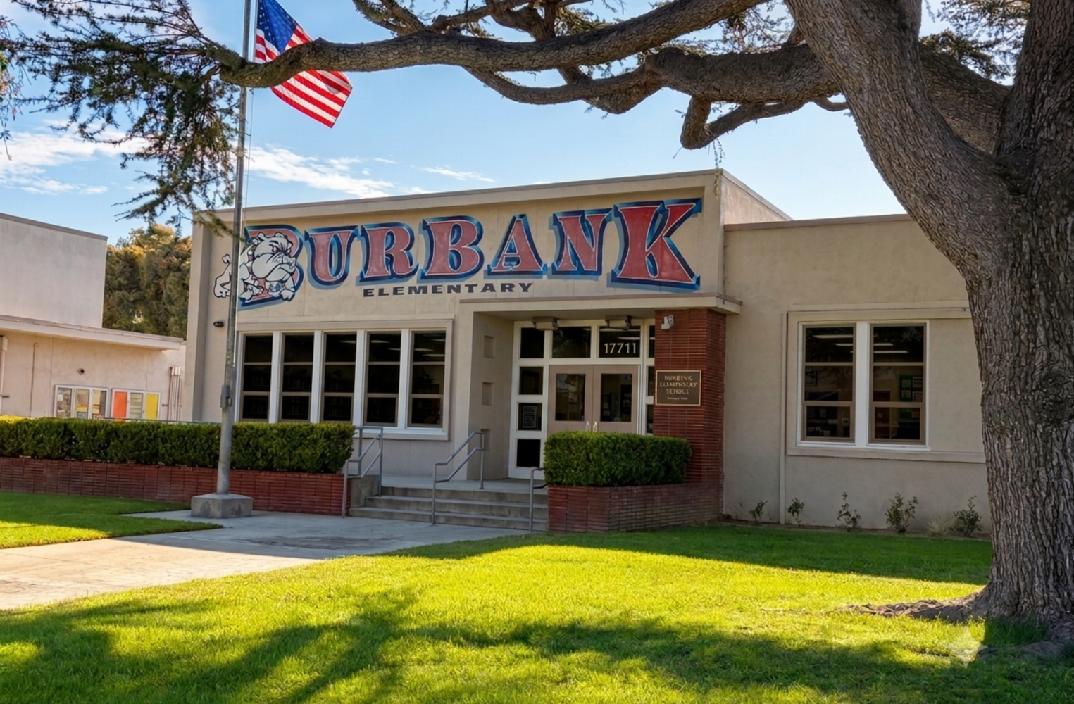Exterior front entrance of Burbank Elementary School featuring red and blue signage, a bulldog mascot logo, an American flag flying on a pole, and a sunny green lawn.