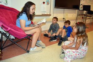 A girl in a chair reads to three children sitting on the floor in a classroom.