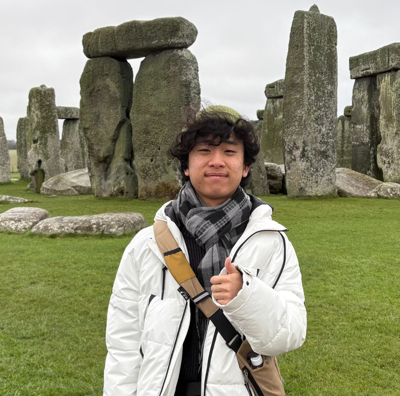 Andrew Kim, a teenage boy with black curly hair wearing a white parka and gray plaid scarf smiling and posing with a thumbs up in front of Stone Henge.