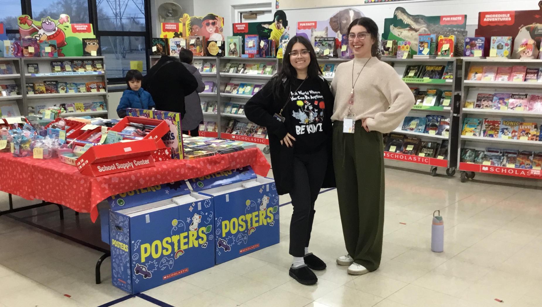 Two women pose in front of a book fair with colorful displays.