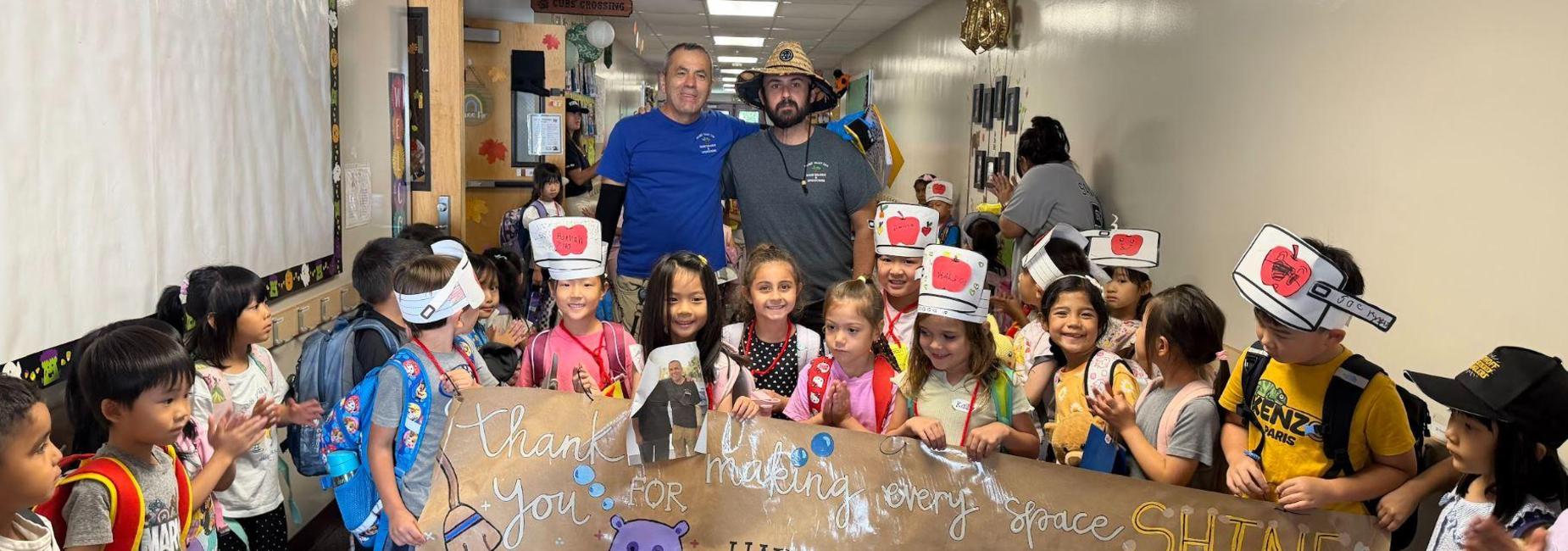 Children and staff holding a thank you banner for custodians in a hallway.
