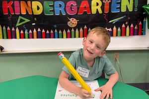 A young student holds a giant pencil to sign his name on a kindergarten welcome sign