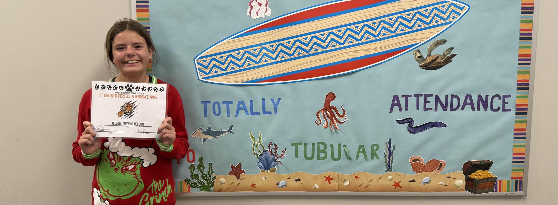A girl in a festive sweater poses with her attendance certificate beside a beach-themed mural.