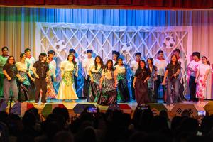 Large group of performers standing and moving in formation on a theater stage under blue, green, and warm lighting. The performers wear a mix of coordinated outfits, including patterned skirts, long skirts, white or dark tops, and casual clothing. A white lattice-style backdrop with large soccer ball decorations spans the stage, with a curtain behind it. Audience silhouettes and raised phones are visible in the foreground.