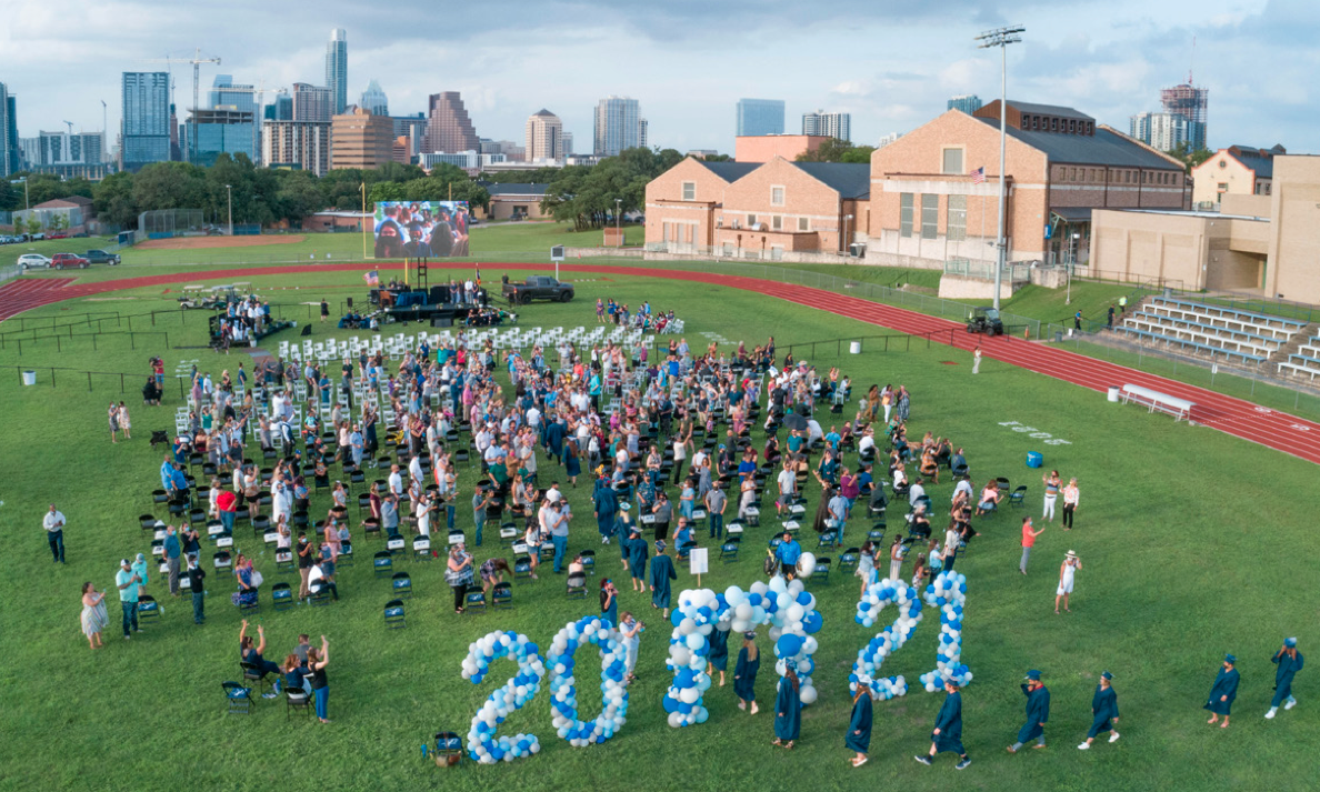 An aerial view of the Texas School for the Deaf 2021 graduation ceremony held on a grassy field with the Austin skyline in the background. Graduates in blue caps and gowns walk among rows of chairs filled with attendees. Large balloon arrangements spell out '2021,' adding a festive touch. A stage and a large video screen display the event, creating a lively and celebratory atmosphere.