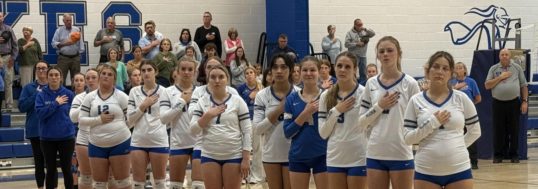 Volleyball players standing in a line with hands on hearts during the national anthem.